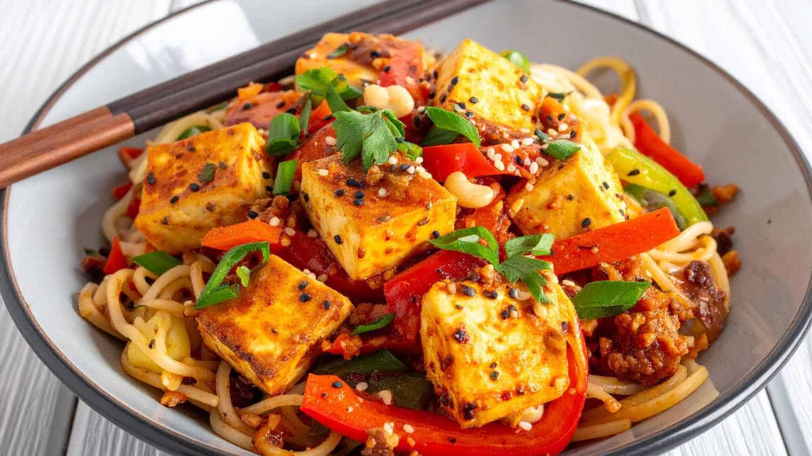 A bowl of noodle stir-fry topped with golden tofu cubes, sliced red peppers, green onions, sesame seeds, and fresh herbs, with chopsticks resting on the bowl’s edge.