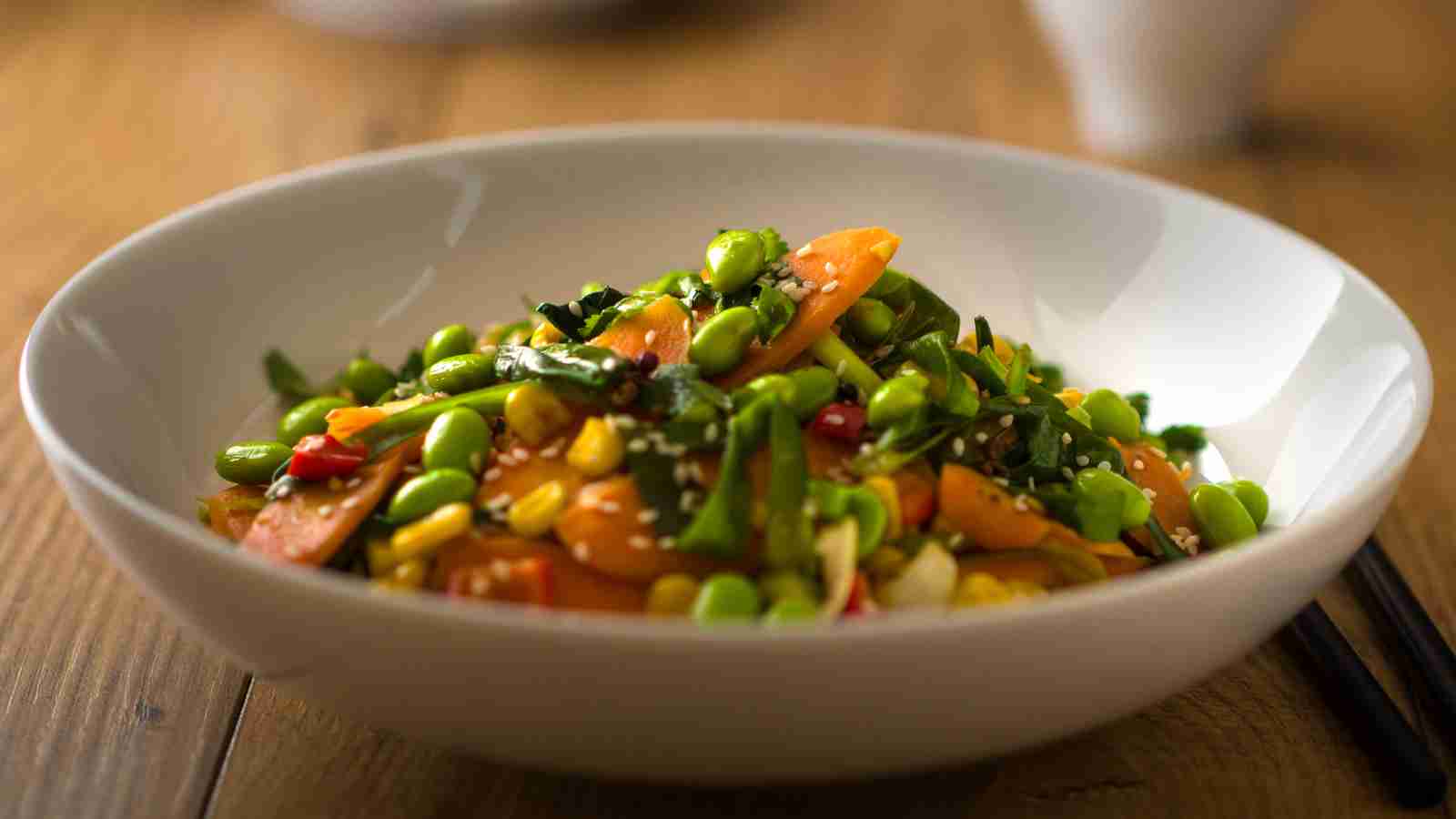 A white bowl filled with a colorful vegetable stir-fry, including edamame, carrots, corn, and chopped greens, sprinkled with sesame seeds.