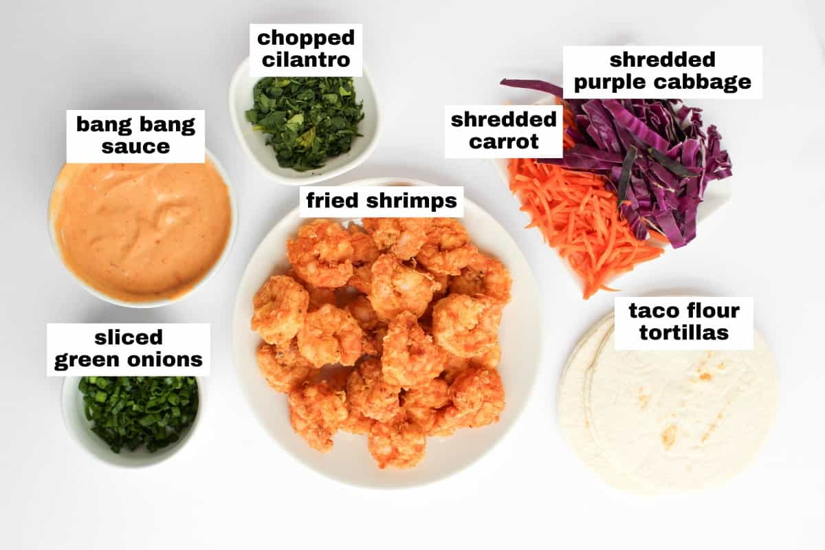A plate of fried shrimps surrounded by bowls of bang bang sauce, sliced green onions, chopped cilantro, shredded purple cabbage, shredded carrot, and taco flour tortillas.
