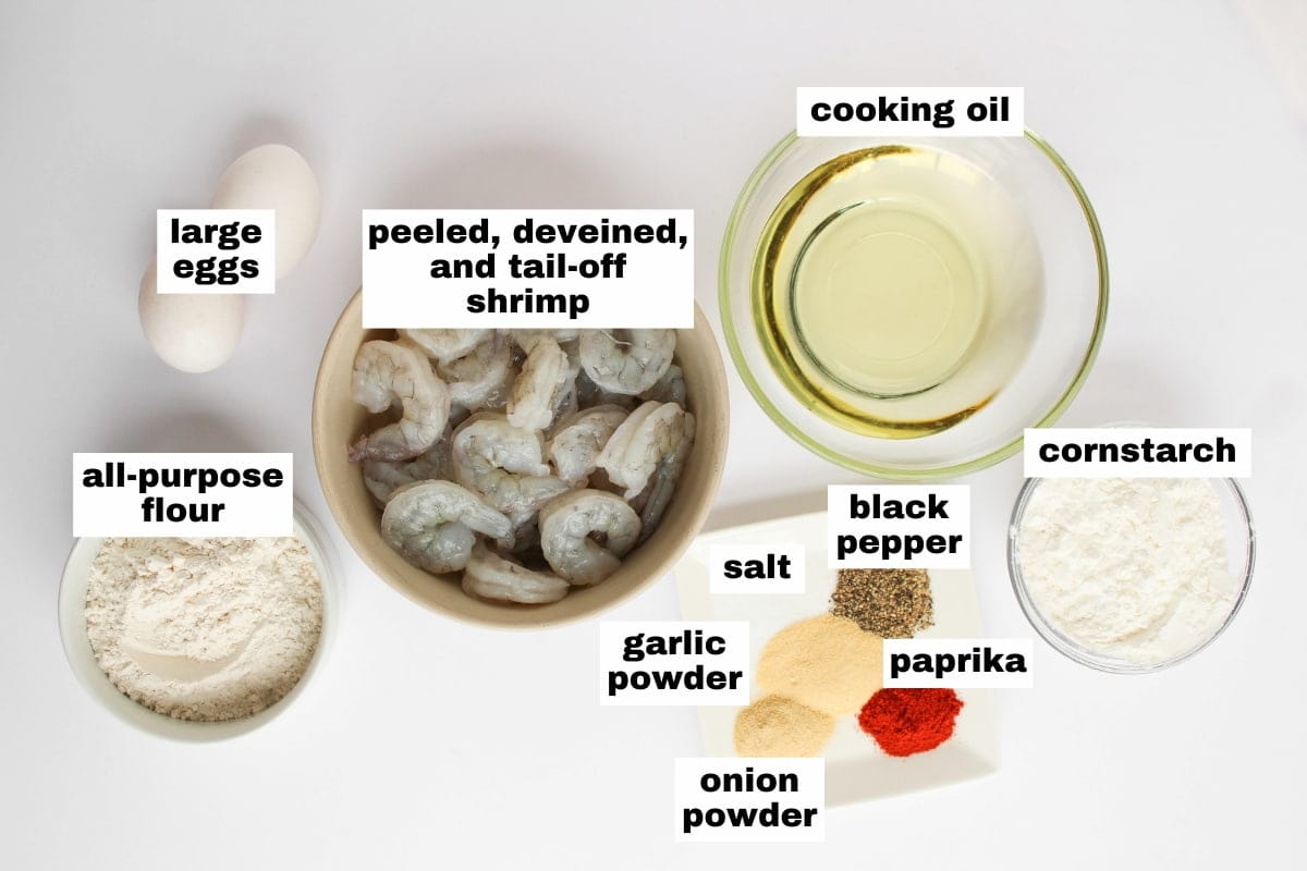 Top view of ingredients in bowls and on a surface: large eggs, shrimp, cooking oil, cornstarch, flour, black pepper, salt, paprika, garlic powder, and onion powder.
