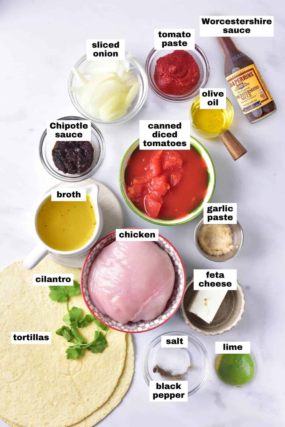 Labeled ingredients for a chicken dish are arranged on a white surface, including chicken, tortillas, canned tomatoes, onion, broth, sauces, feta, lime, and seasonings.