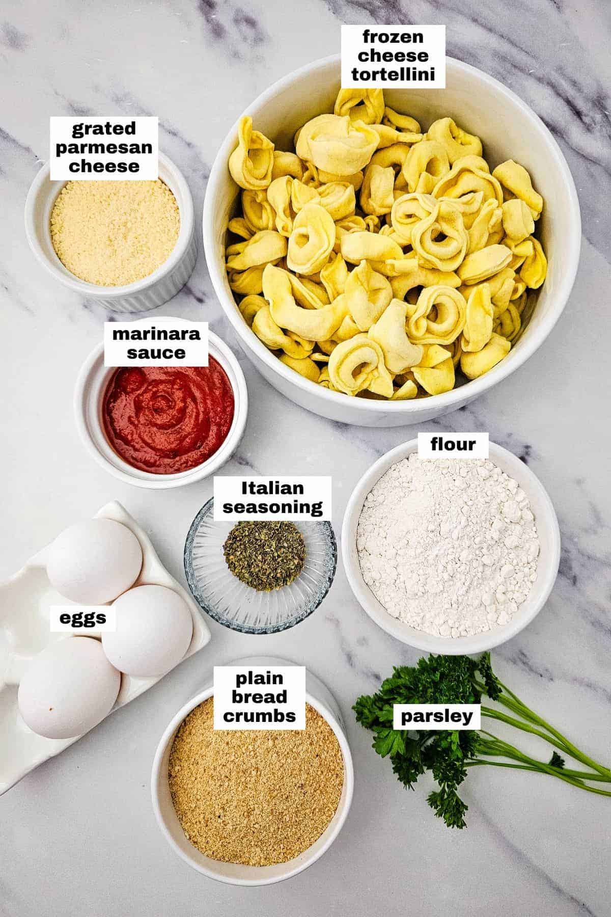 Bowls and plates containing frozen cheese tortellini, flour, eggs, plain bread crumbs, grated parmesan, marinara sauce, Italian seasoning, and parsley on a marble surface.