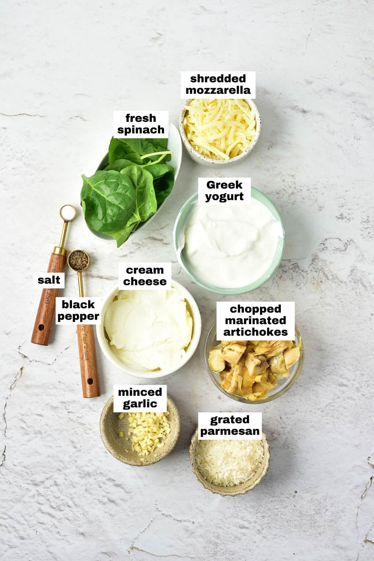 Ingredients for a recipe arranged on a white surface, including fresh spinach, mozzarella, Greek yogurt, cream cheese, marinated artichokes, parmesan, minced garlic, salt, and black pepper.