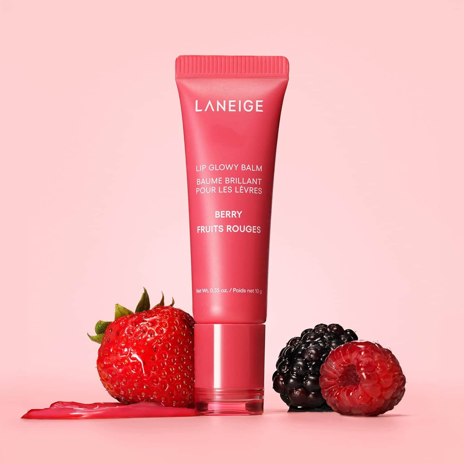 Laneige Lip Glowy Balm in Berry flavor stands upright on a pink surface, surrounded by a strawberry, blackberry, raspberry, and a smear of pink balm.
