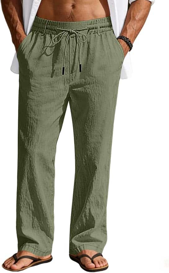 A person wearing light olive green drawstring linen pants, a white shirt, a beaded bracelet, and black flip-flops, standing with hands in pockets.