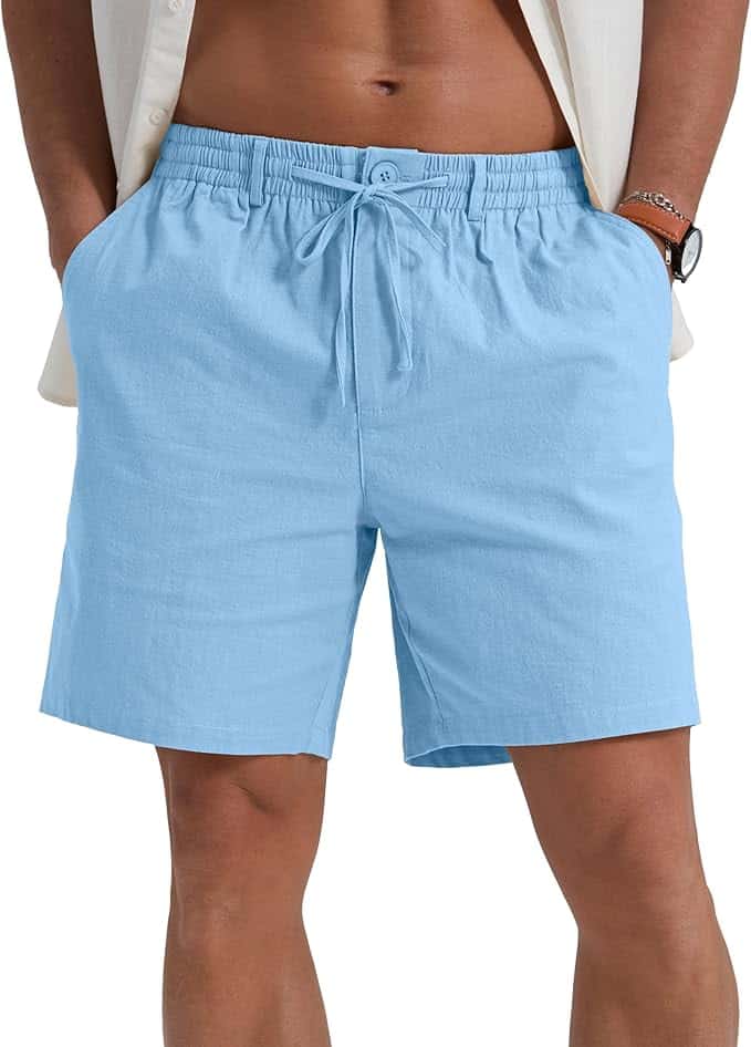 A person wearing light blue drawstring shorts with hands in pockets, white shirt open over chest, and a watch on left wrist.
