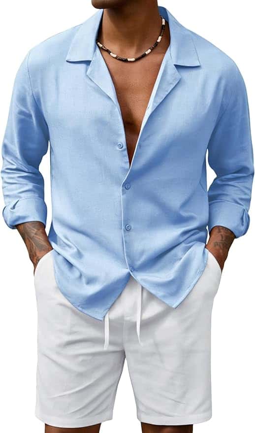 Man wearing a light blue button-up shirt with rolled-up sleeves, white shorts, and a beaded necklace, standing with hands in pockets.
