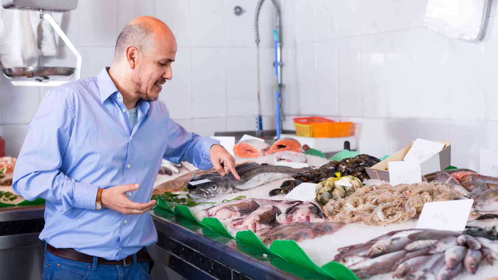 A man in a blue shirt examines fresh fish and seafood displayed on ice at a market counter.