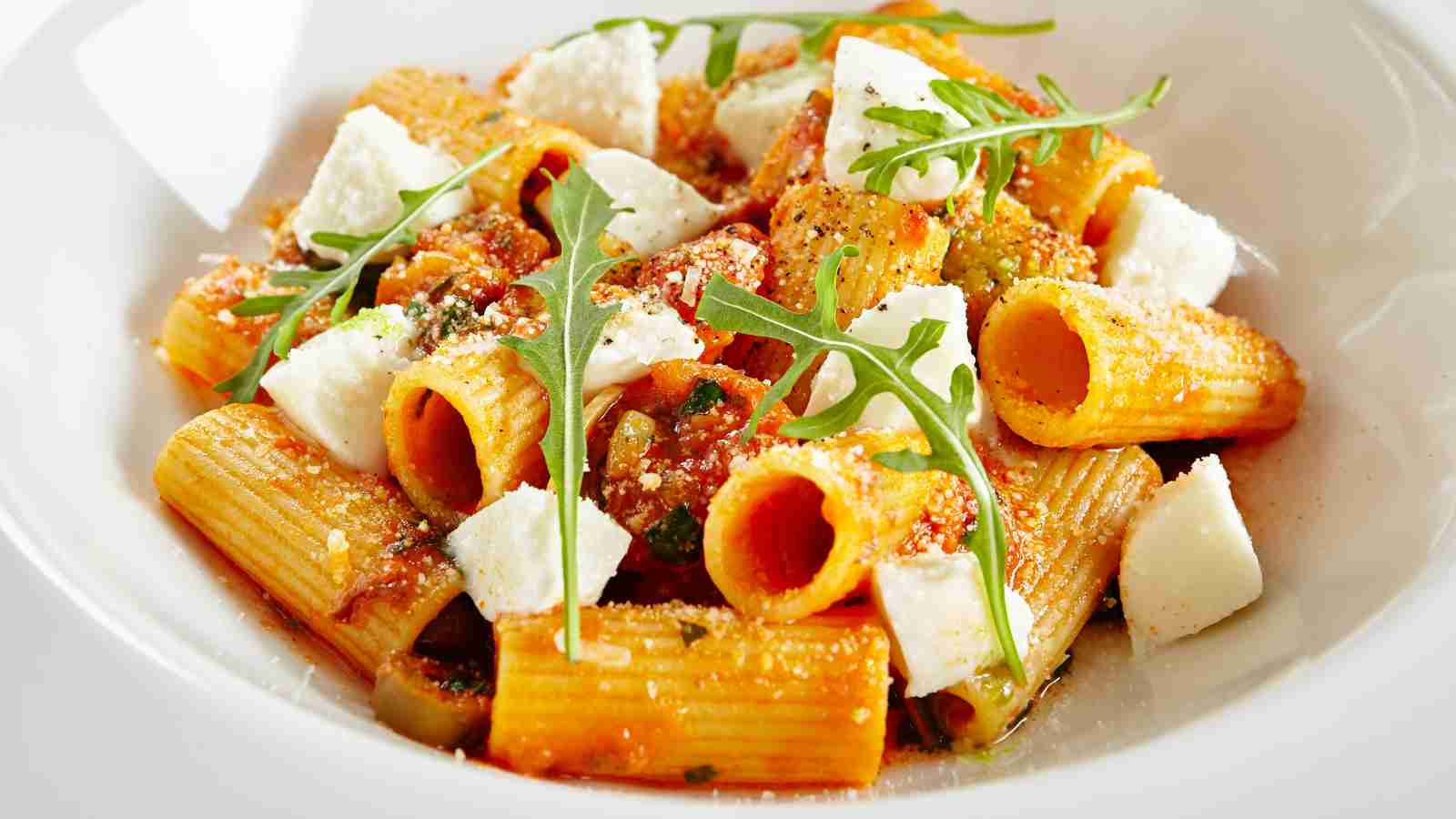 A plate of rigatoni pasta with tomato sauce, chunks of white cheese, grated parmesan, and arugula leaves, served in a white bowl.