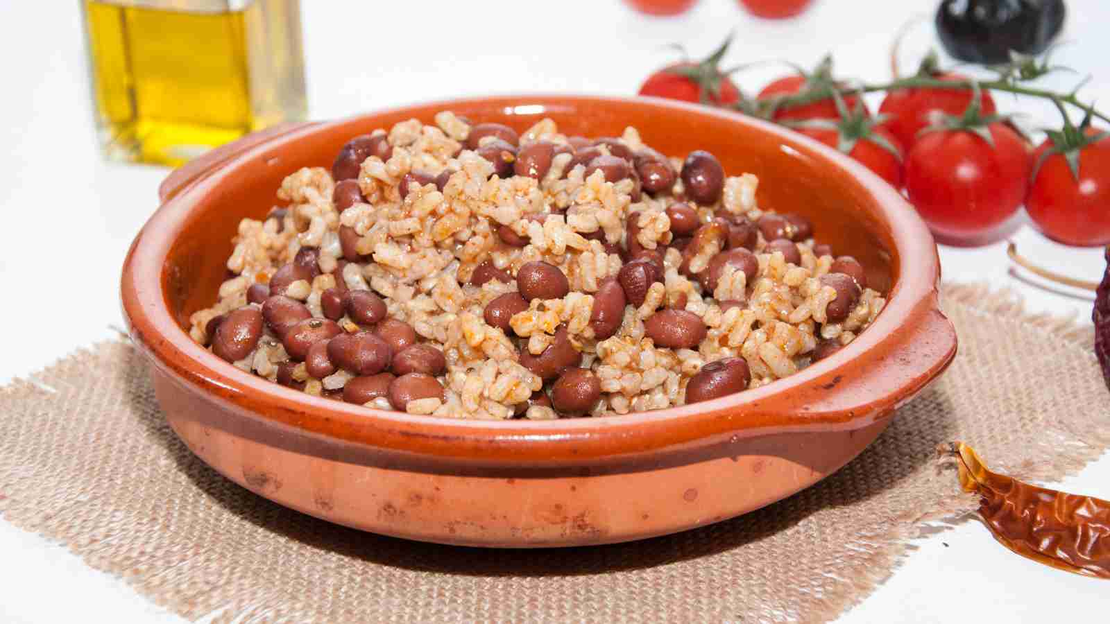 A brown ceramic bowl filled with cooked rice and beans sits on a burlap mat, with tomatoes and a bottle of oil in the background.