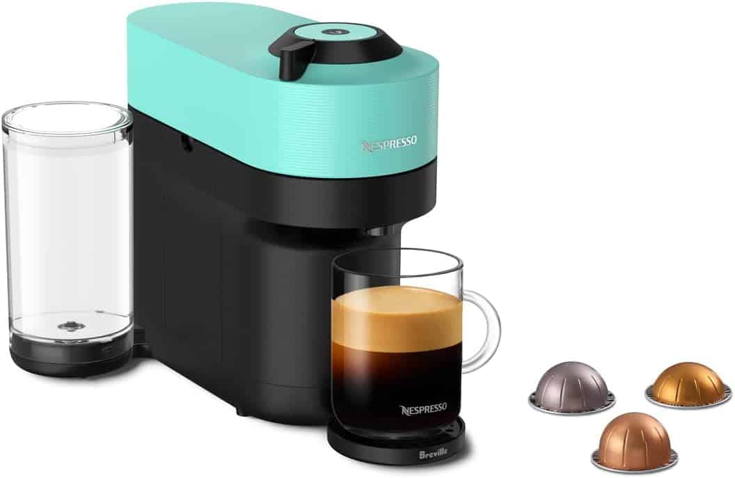 A teal and black Nespresso coffee machine with a glass mug of coffee, water reservoir, and four assorted coffee pods placed beside it.