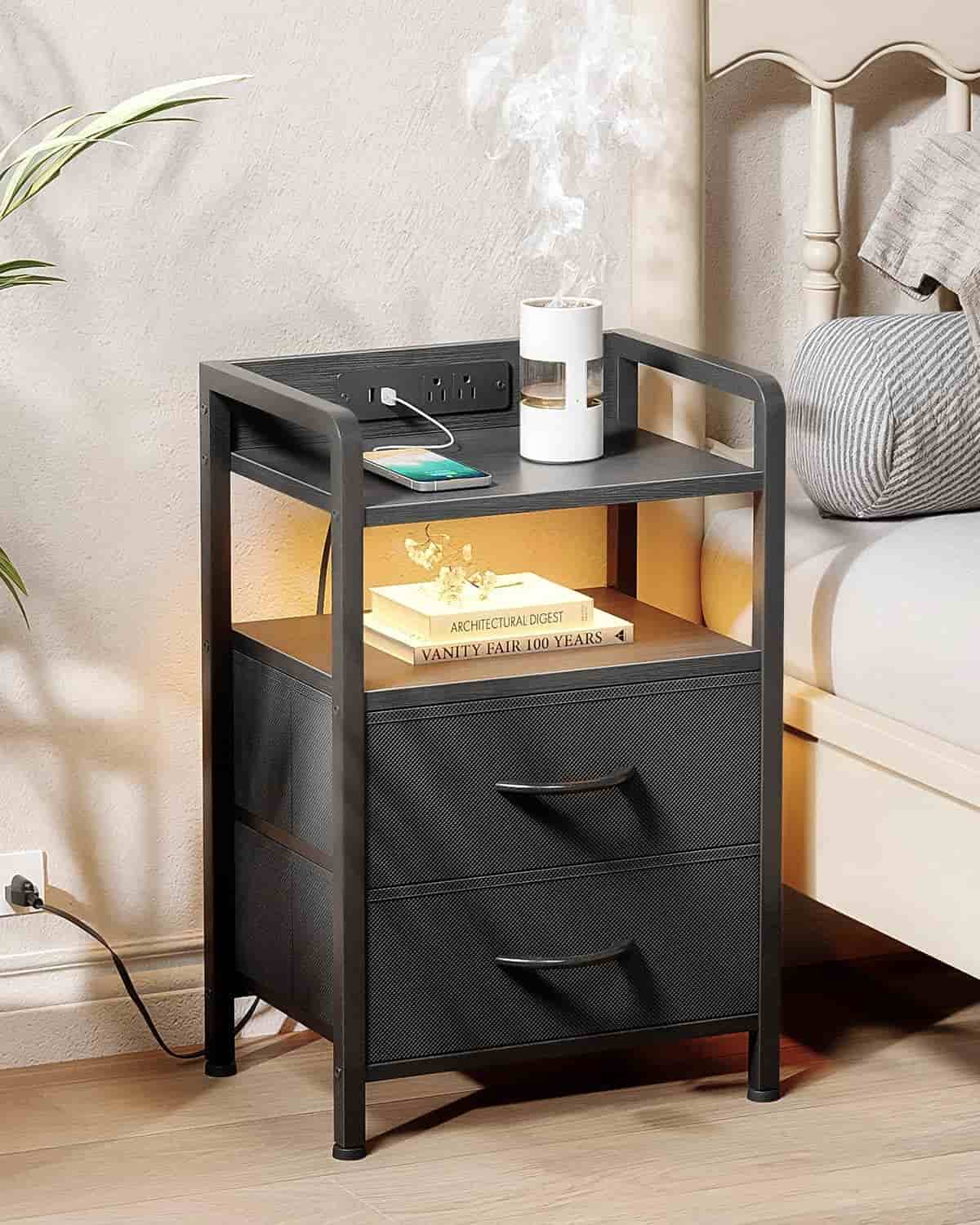 Black metal and wood nightstand with two drawers, a shelf holding books, a phone charging on top, and a small lamp; electrical outlet built into the back.