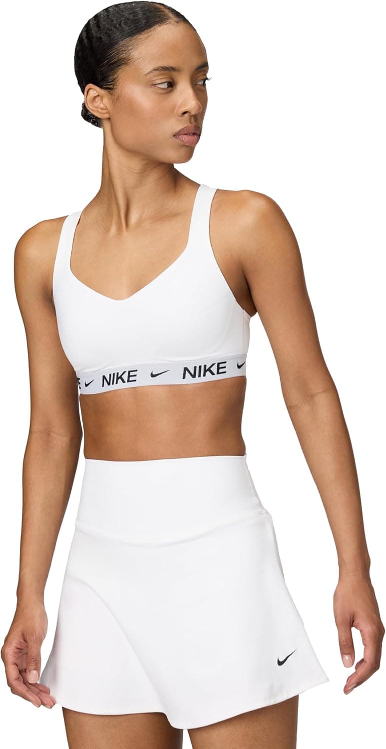 A woman in a white sports bra with "NIKE" branding and a matching white athletic skirt stands against a plain background, looking to her left.