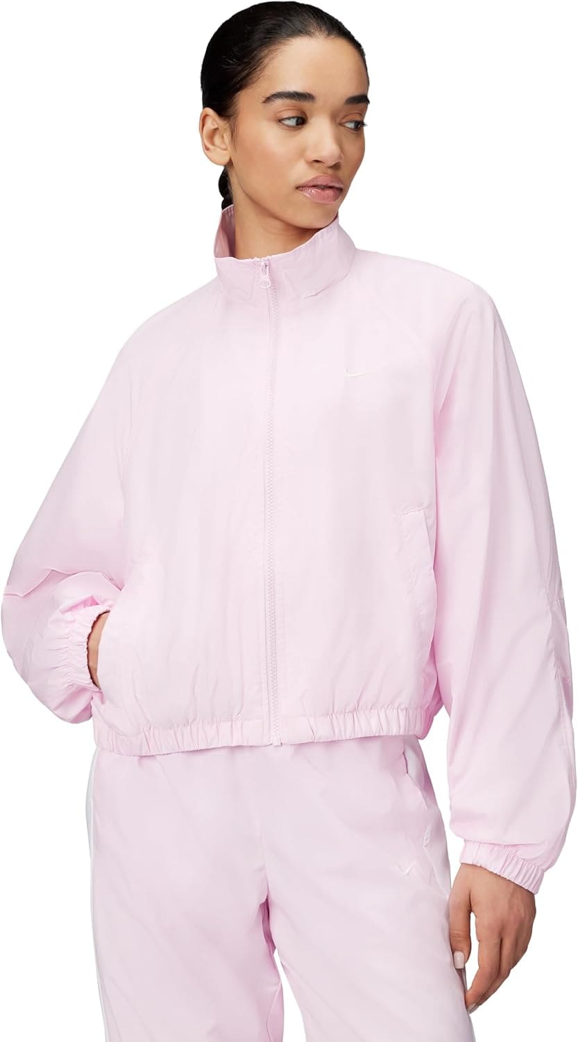 A woman wearing a light pink zip-up jacket and matching pants stands with one hand in her pocket, looking to the side.