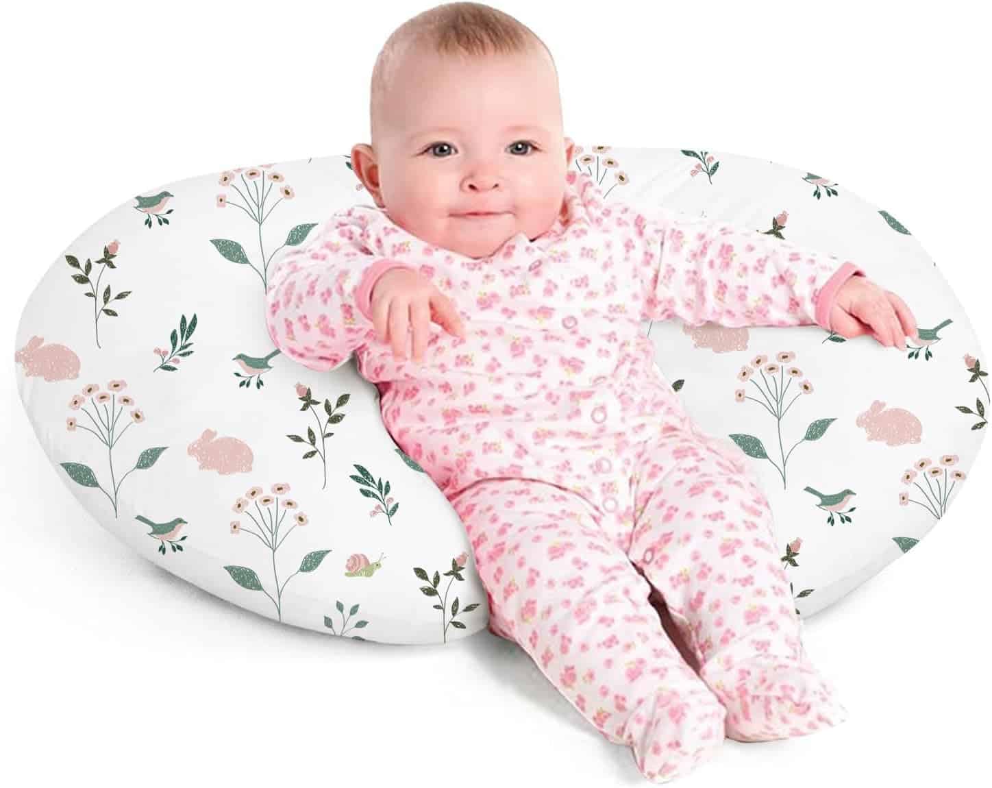 A baby wearing a pink patterned onesie sits on a white, oval-shaped pillow with a floral and bunny print.