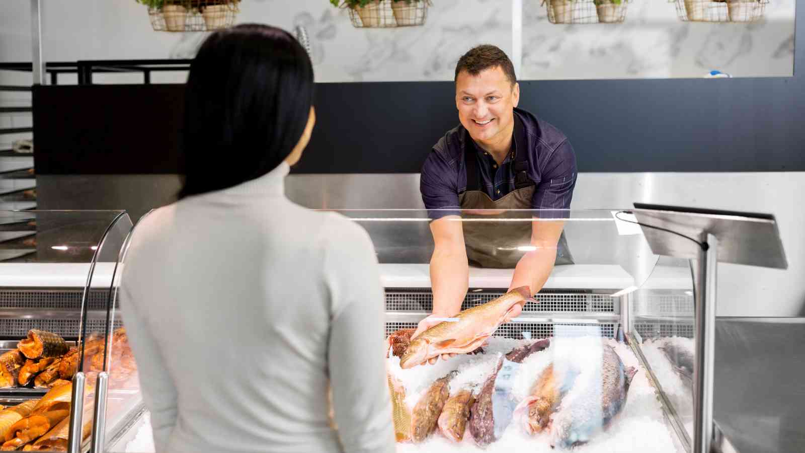 A fishmonger behind a counter hands a whole fish to a customer. Various fish and seafood are displayed on ice in the display case.