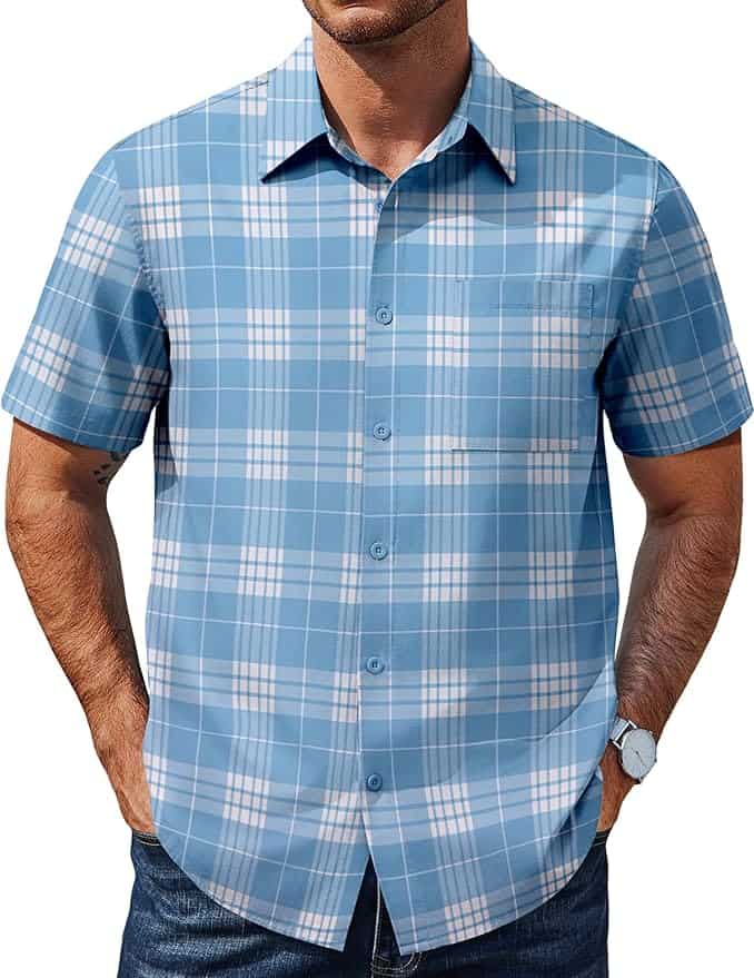 A man wearing a short-sleeve, button-up blue plaid shirt with white and light blue patterns, standing with hands in his pockets.