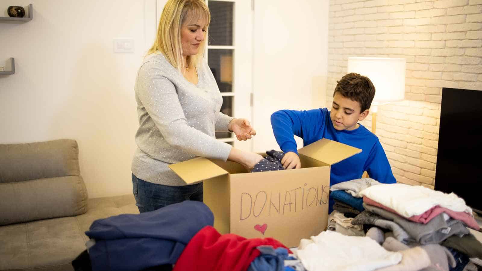 A woman and a boy place folded clothes into a cardboard box labeled "Donations" in a living room with a pile of clothes nearby.