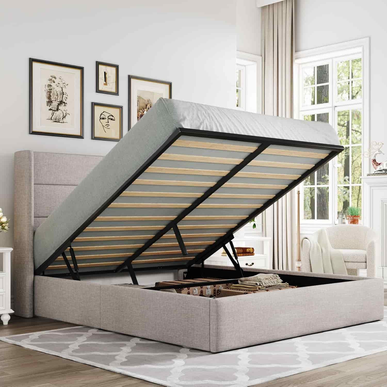 A modern upholstered bed with a lifted mattress frame revealing storage space underneath, set in a bright bedroom with framed art and large windows.