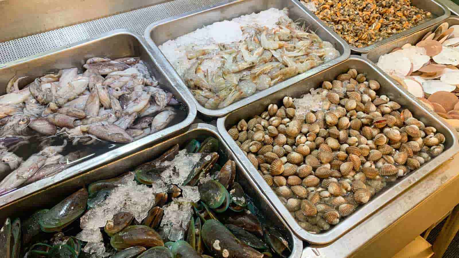 Assorted fresh seafood, including shrimp, mussels, clams, and squid, displayed on ice in metal trays at a market.