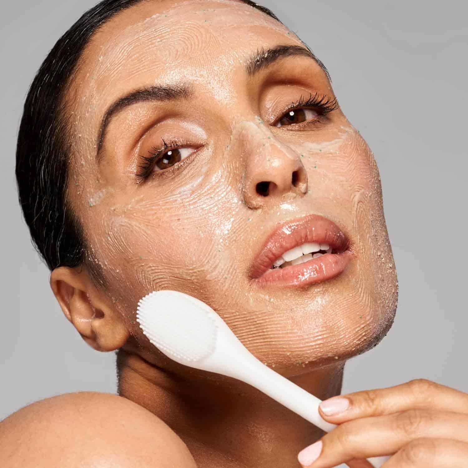 A woman applies a facial cleanser or mask with a textured silicone brush on her face, showcasing skincare routine.