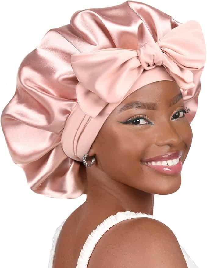 Woman smiling and wearing a shiny pink satin bonnet with a large bow, dressed in a white top, shown against a plain background.