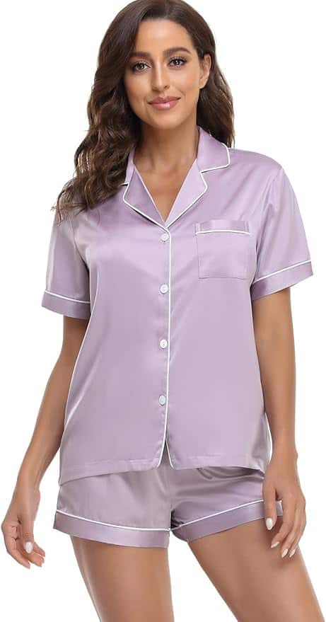 Woman wearing a short-sleeve, button-up satin pajama set in light lavender with white piping, featuring a chest pocket and matching shorts.