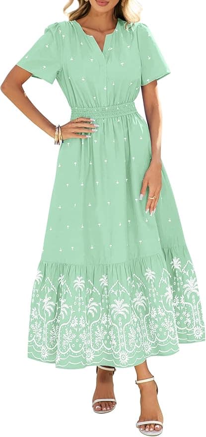 Woman wearing a light green, short-sleeved maxi dress with white embroidered patterns near the hem, standing in sandals with one hand on her hip.