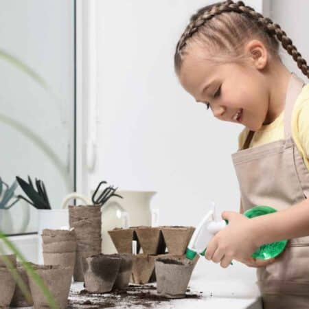 A young girl wearing an apron waters small plant pots with a spray bottle on an indoor table, surrounded by gardening tools and soil.