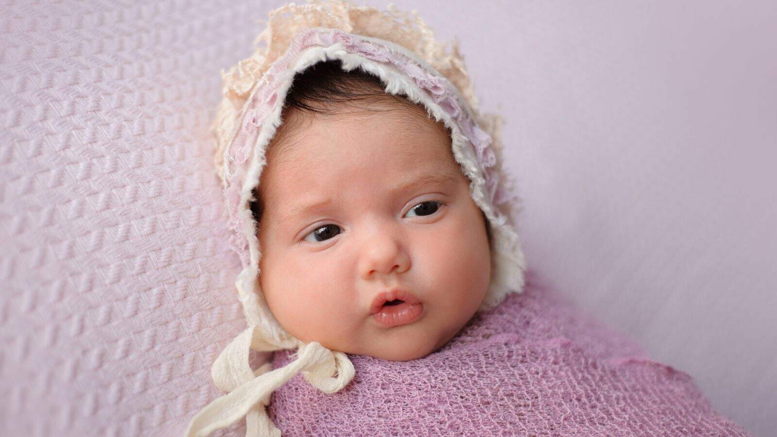 A baby wrapped in a light purple blanket and wearing a lace bonnet, lying on a textured, light purple surface.