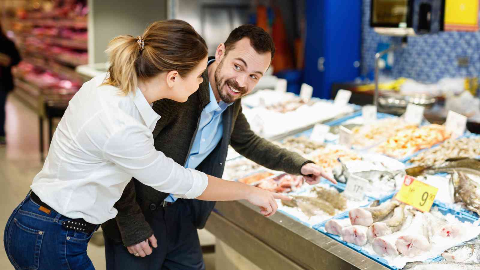 Two people stand at a seafood counter in a grocery store, looking at and pointing to various fish and seafood displayed on ice.