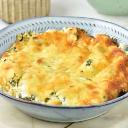 A baked dish with a golden-brown cheese topping is served in a patterned bowl, placed on a round white board.