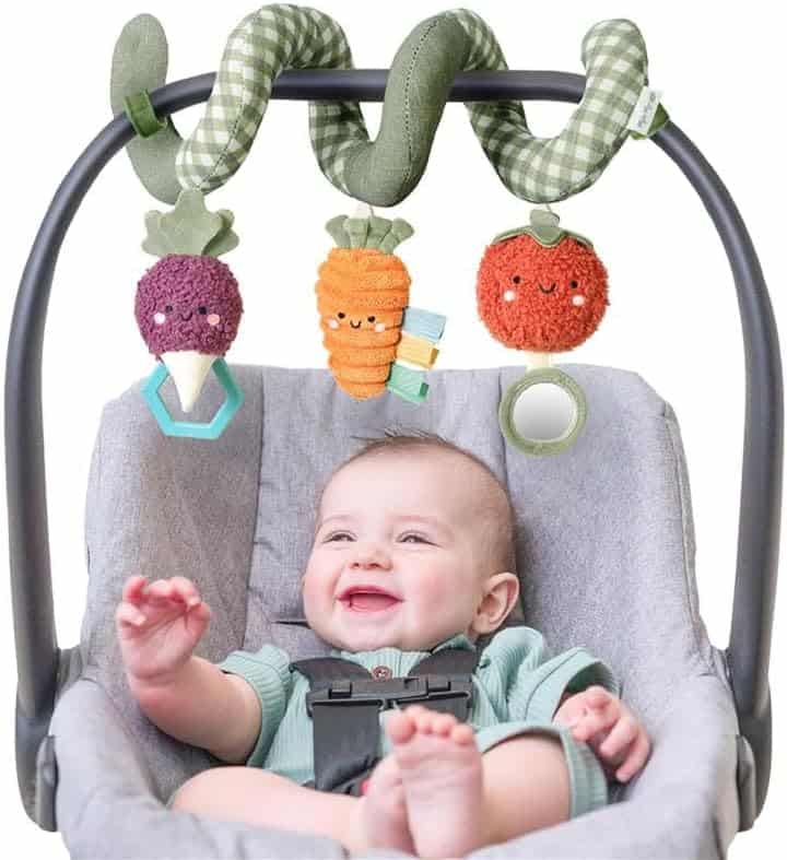 A smiling baby in a car seat reaches toward a soft spiral toy with plush vegetable figures hanging above.
