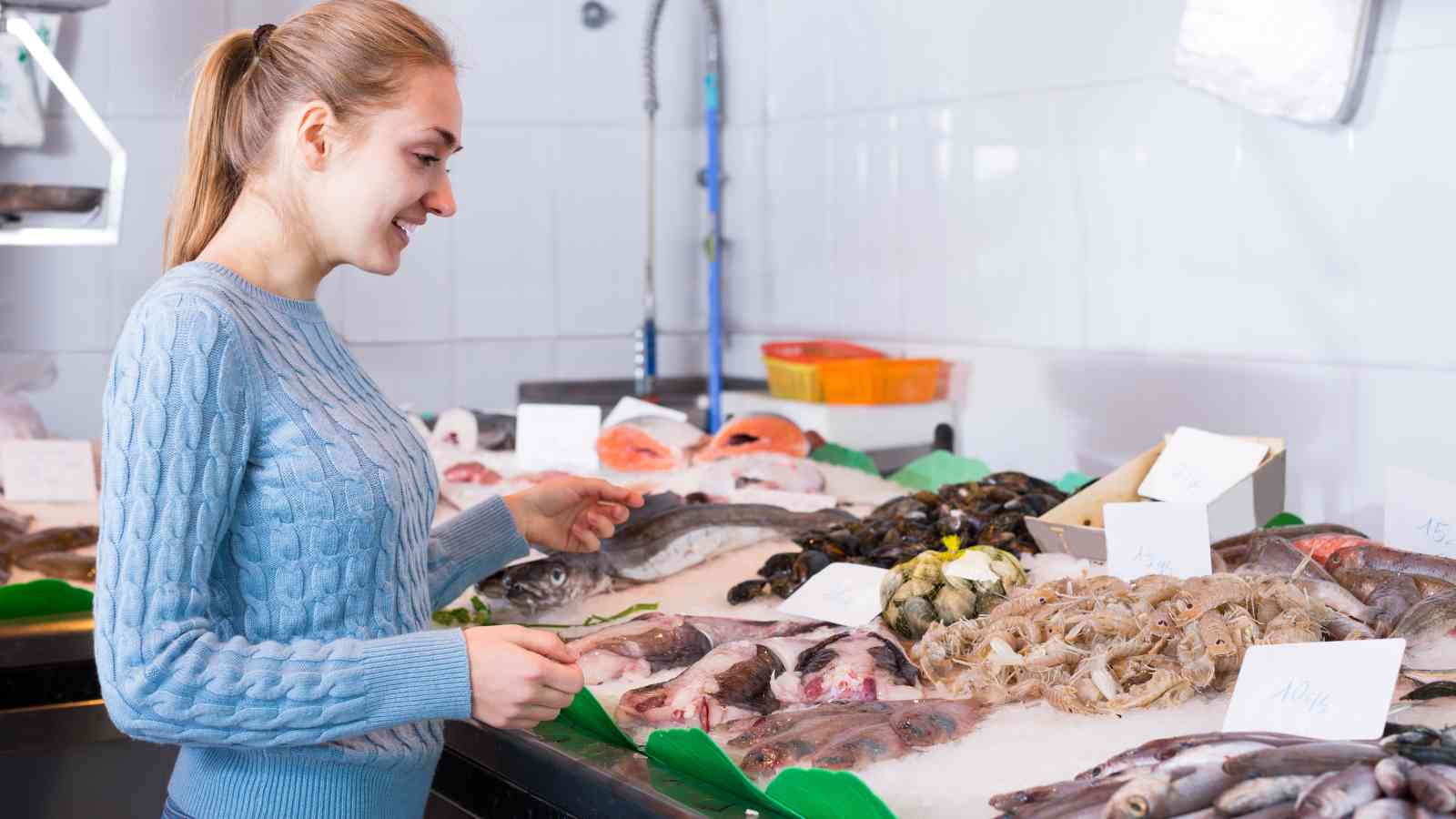 A woman in a blue sweater selects fresh seafood from a display at a fish market, with various fish, shrimp, and shellfish on ice.