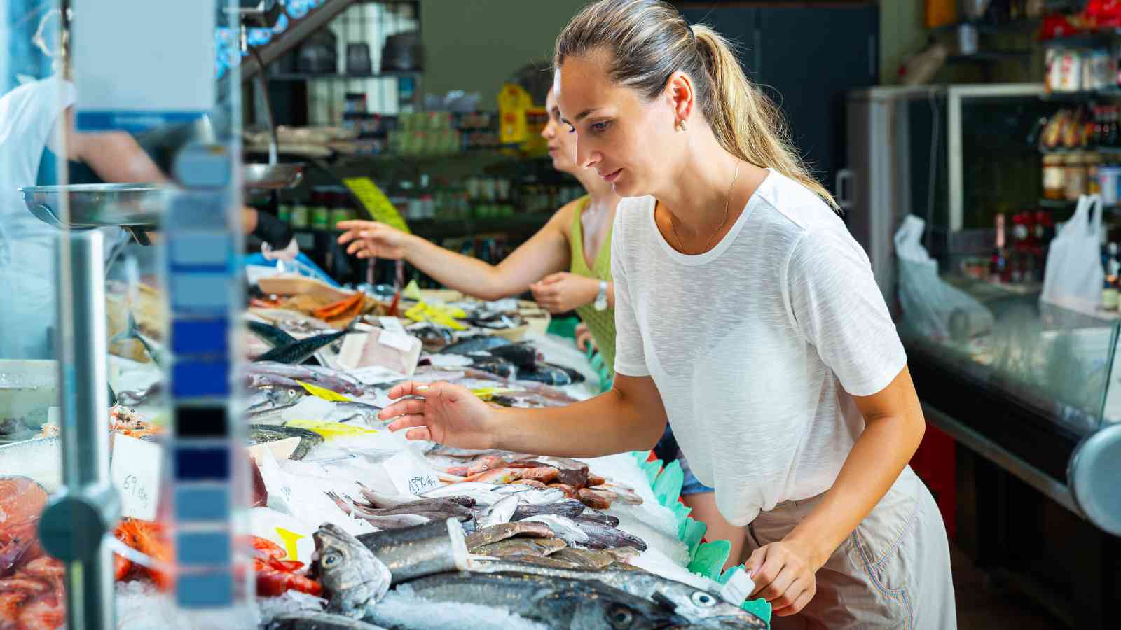 A woman examines various types of fresh fish and seafood displayed on ice at a market counter.