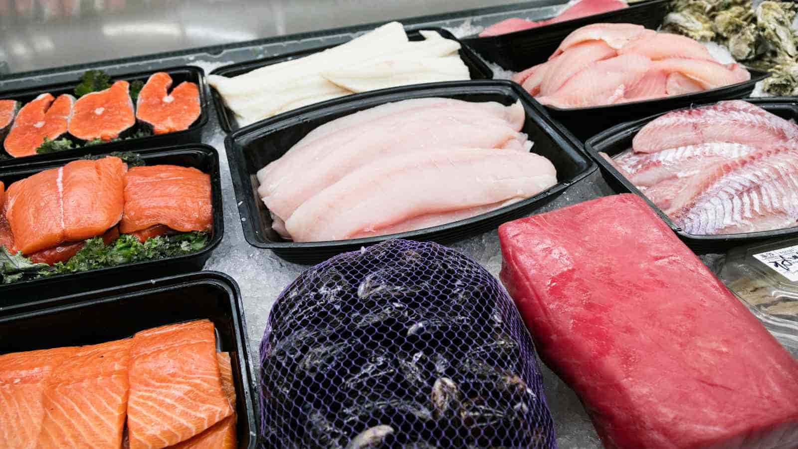 Variety of raw seafood including salmon, white fish fillets, tuna, and shellfish displayed in trays on ice at a seafood market.