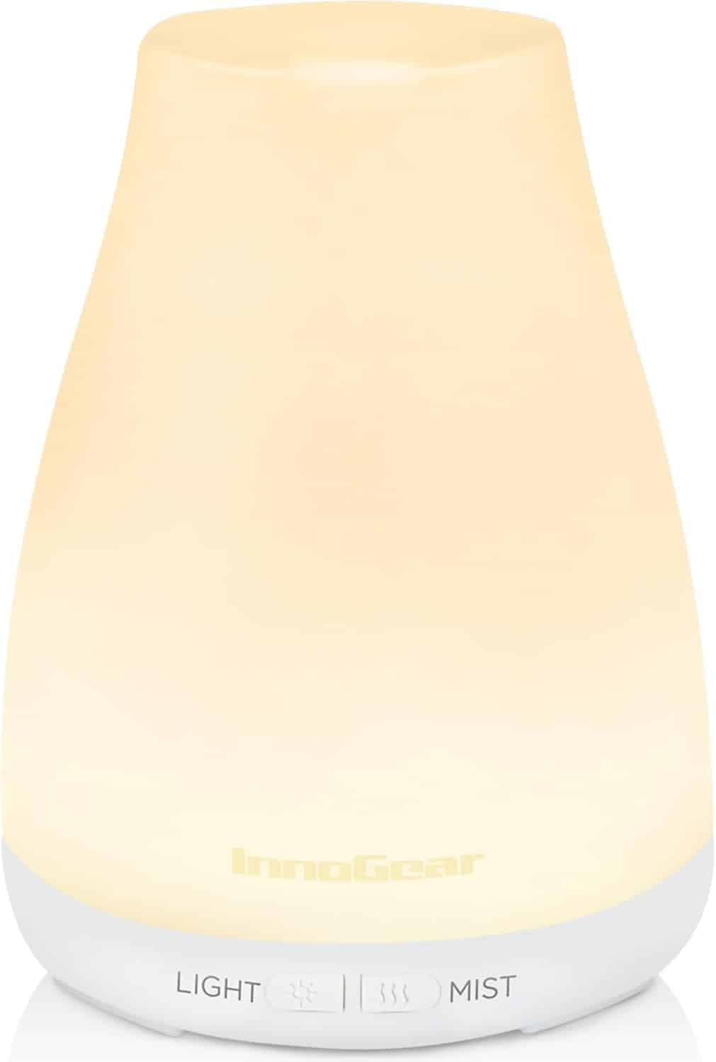 A lit InnoGear essential oil diffuser with a white base and glowing yellow light, featuring labeled buttons for light and mist settings at the bottom.
