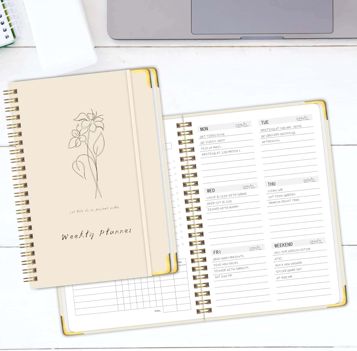 An open spiral-bound weekly planner displays a week’s schedule, with another closed planner on top, both placed on a white desk near a laptop.