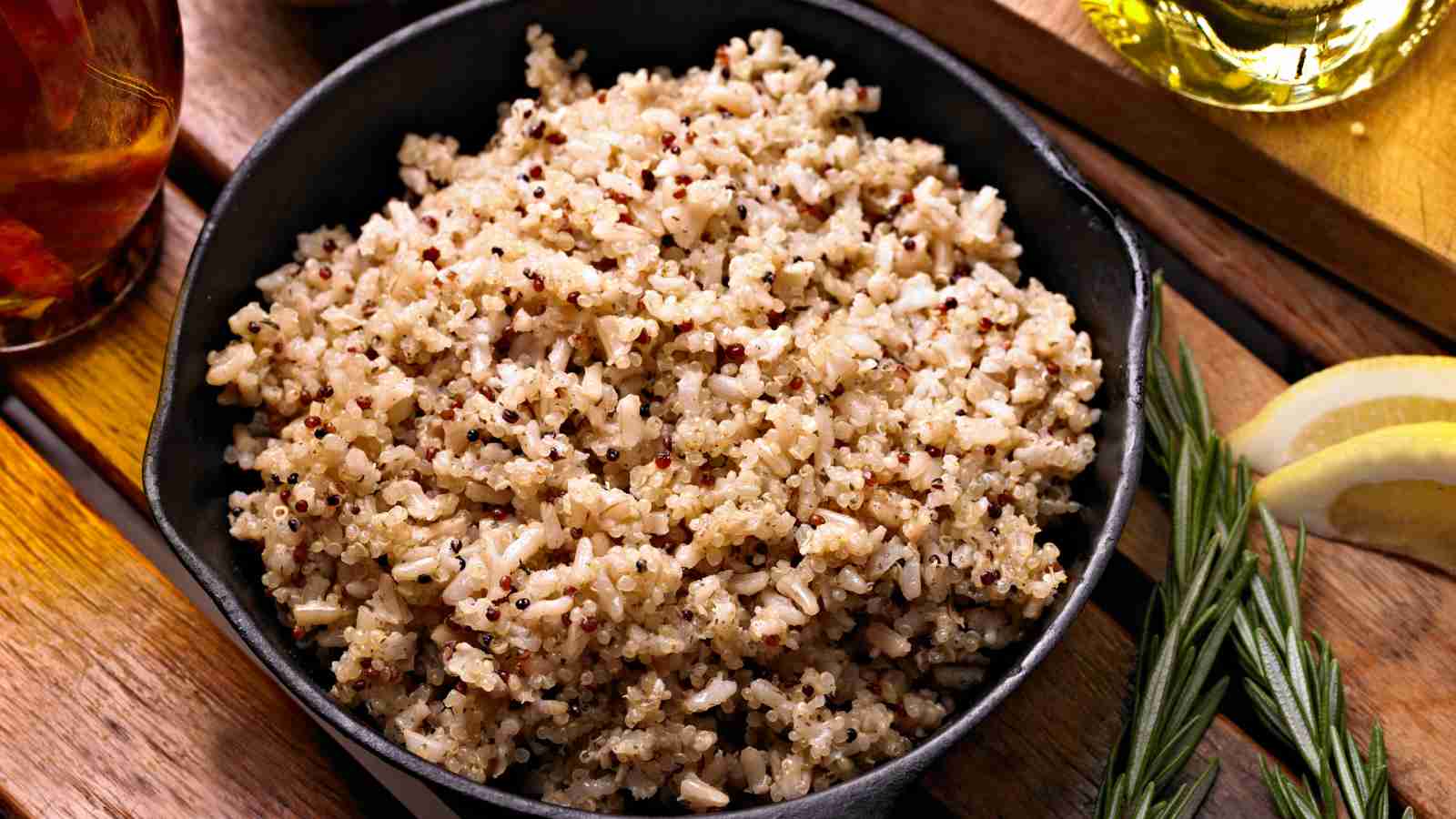 A bowl of cooked quinoa and brown rice blend sits on a wooden table, with rosemary, lemon slices, and a glass of liquid nearby.