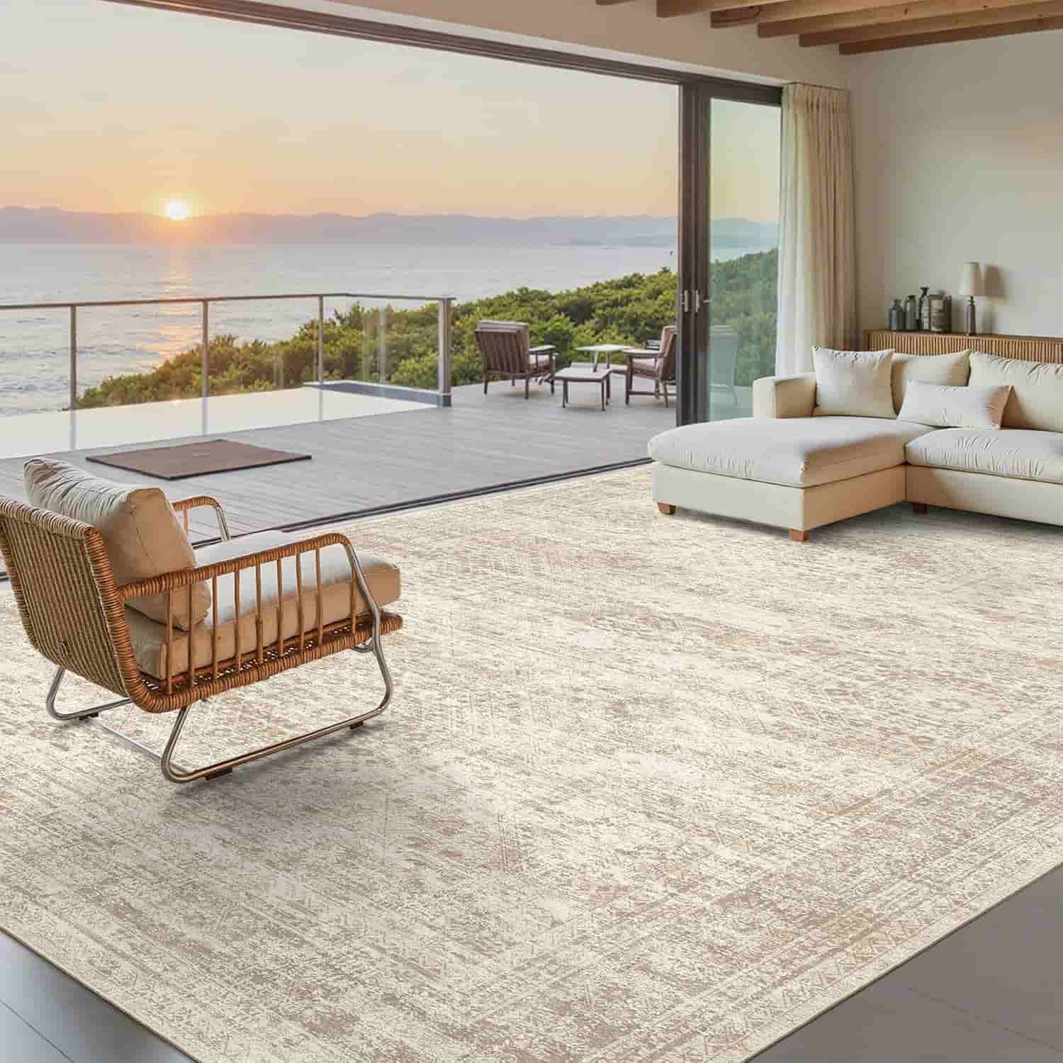 Modern living room with a large cream-colored rug, cushioned sectional sofa, wicker chair, and glass doors opening to a deck with ocean and sunset view.