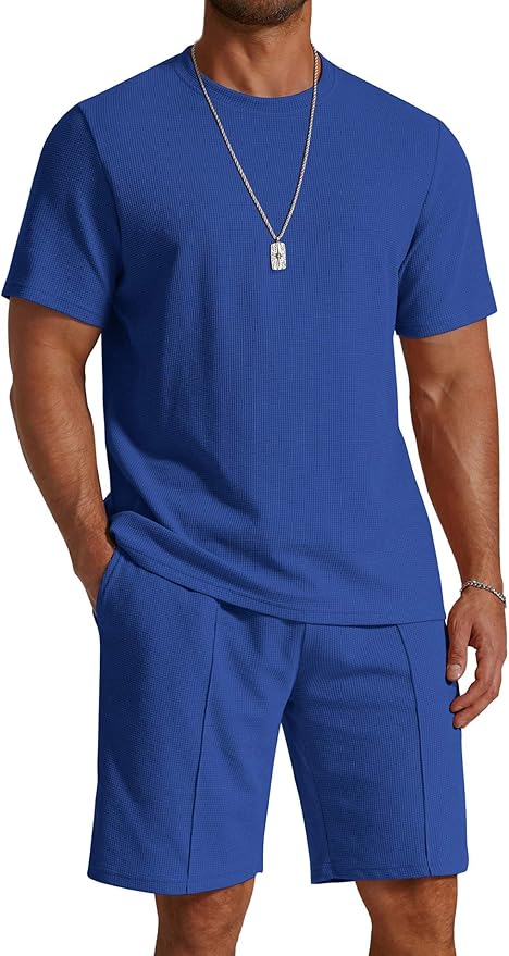 Man wearing a matching royal blue short-sleeve shirt and shorts set with a silver necklace, standing with one hand in his pocket.