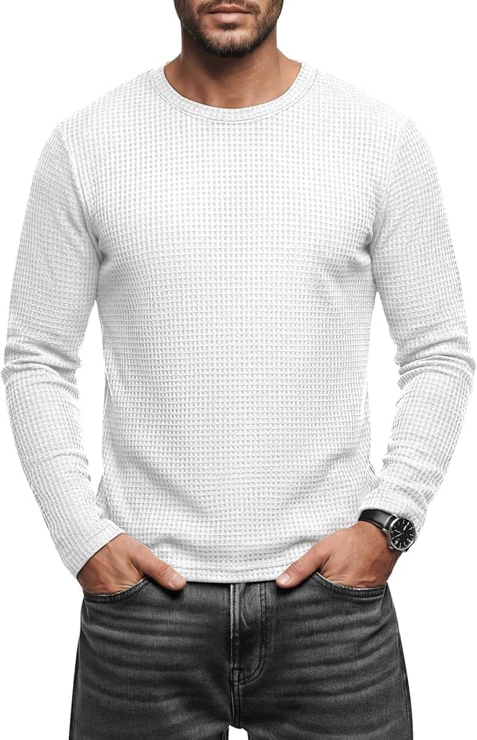 A man wearing a white textured long-sleeve shirt and dark jeans stands with his hands in his pockets, facing forward.