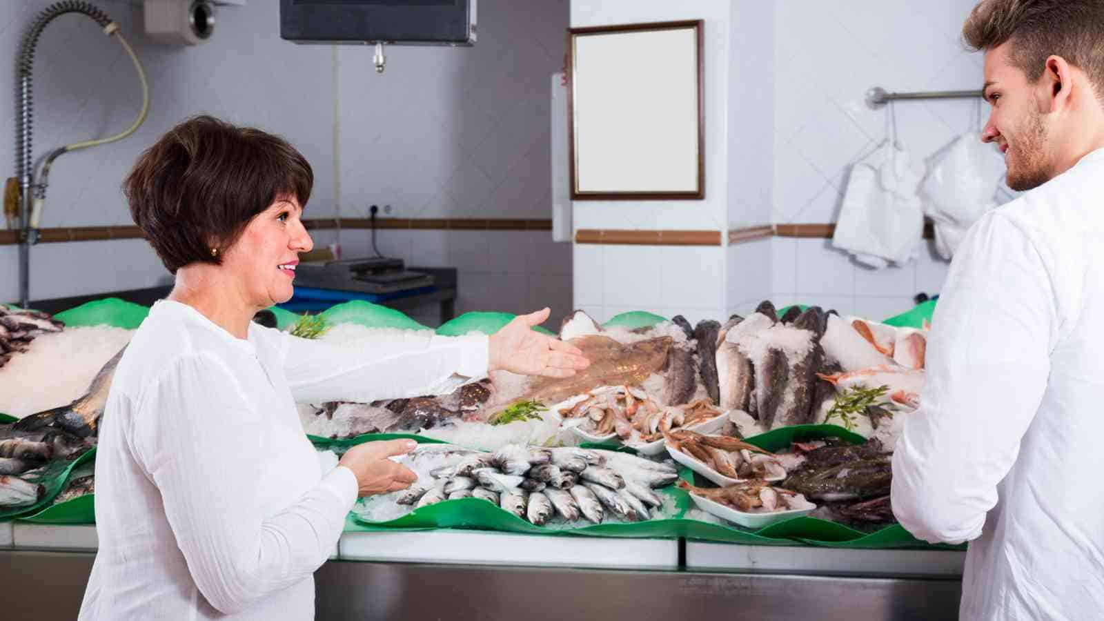 A woman and a man talk in front of a fish counter displaying various fresh seafood in a market setting.