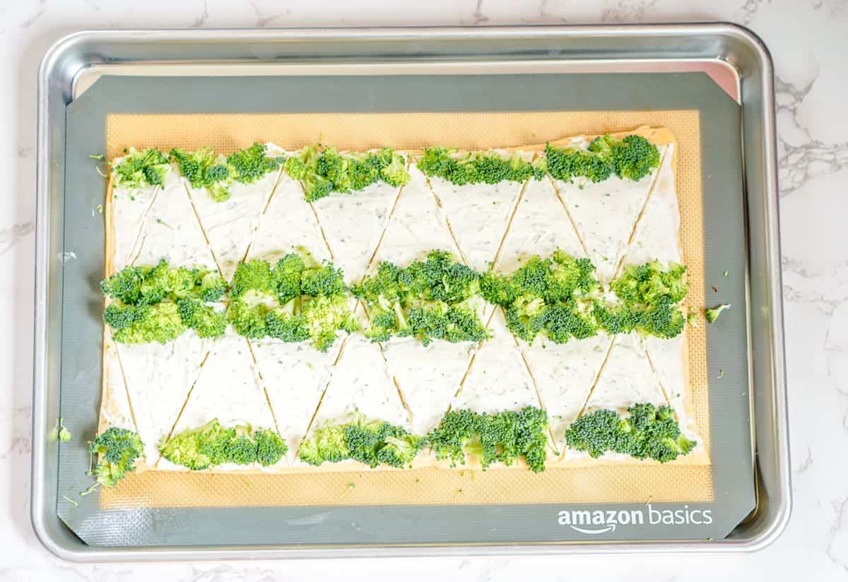 A baking sheet with triangular pieces of dough topped with a creamy white spread and fresh broccoli florets, arranged in rows.