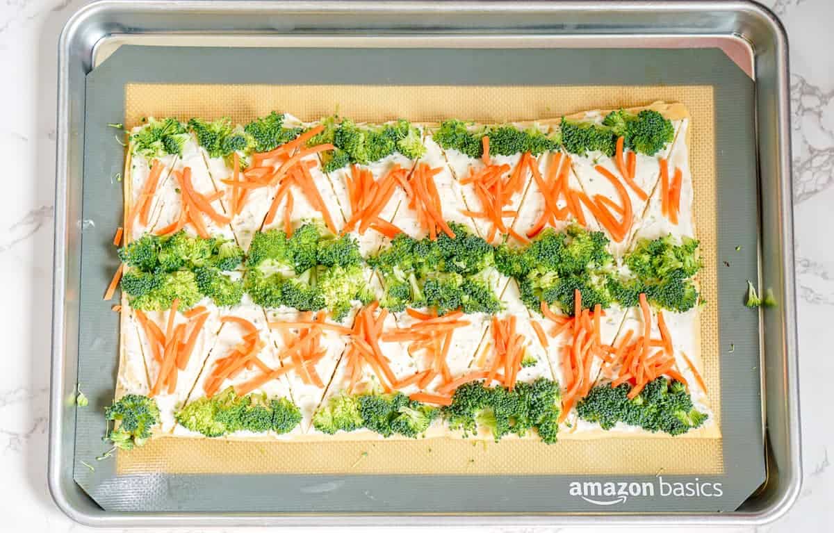 Rectangular sheet of dough topped with cream cheese, shredded carrots, and broccoli florets on a baking tray with an Amazon Basics silicone mat.
