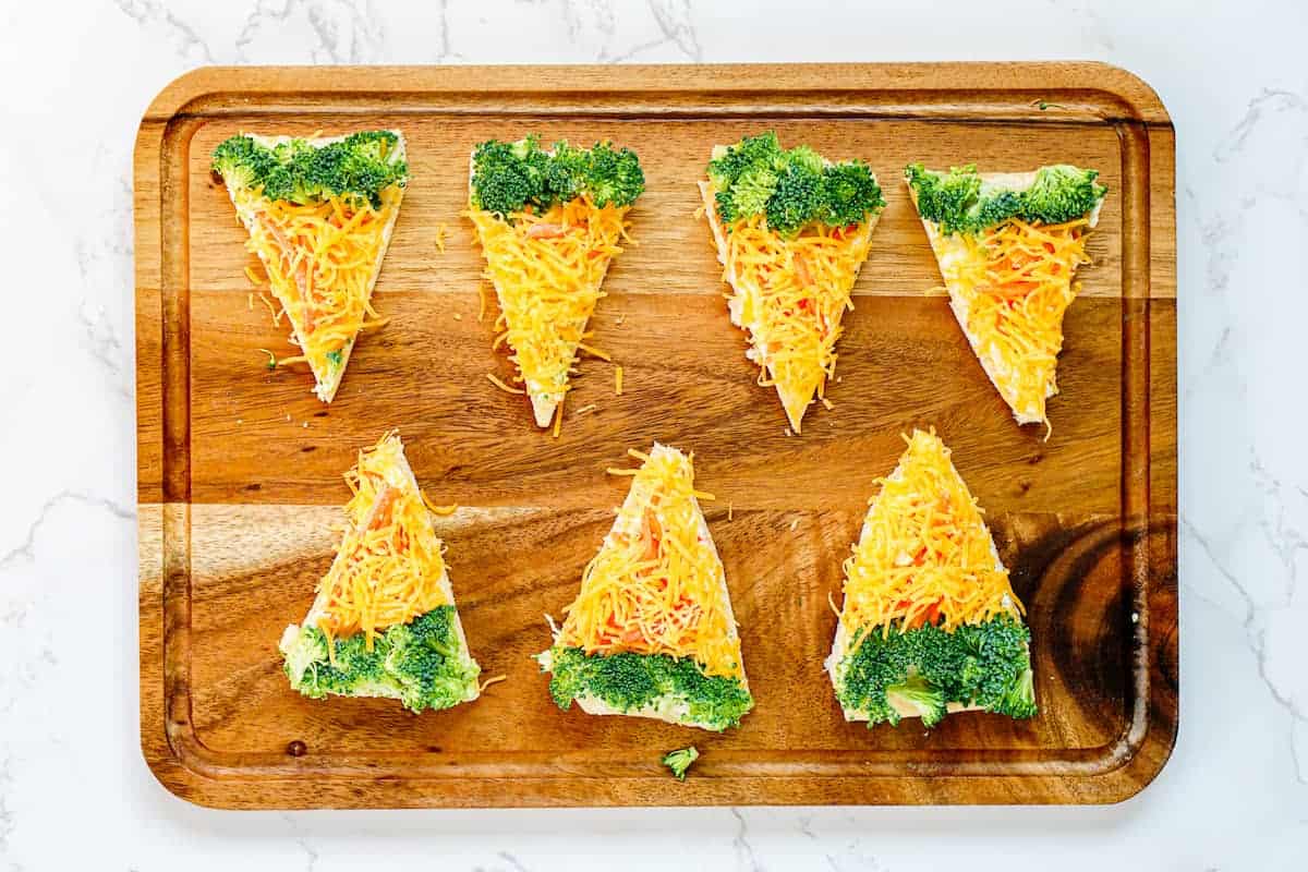 Six triangle-shaped pieces of bread topped with shredded cheese and broccoli, arranged on a wooden cutting board.