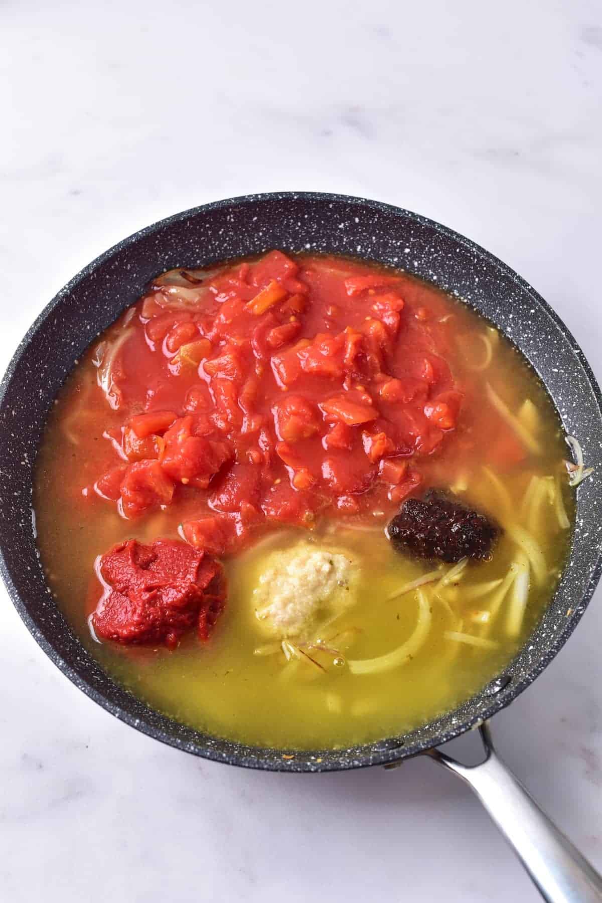 A frying pan containing diced tomatoes, tomato paste, grated garlic, a dark paste, sliced onions, and broth on a white surface.