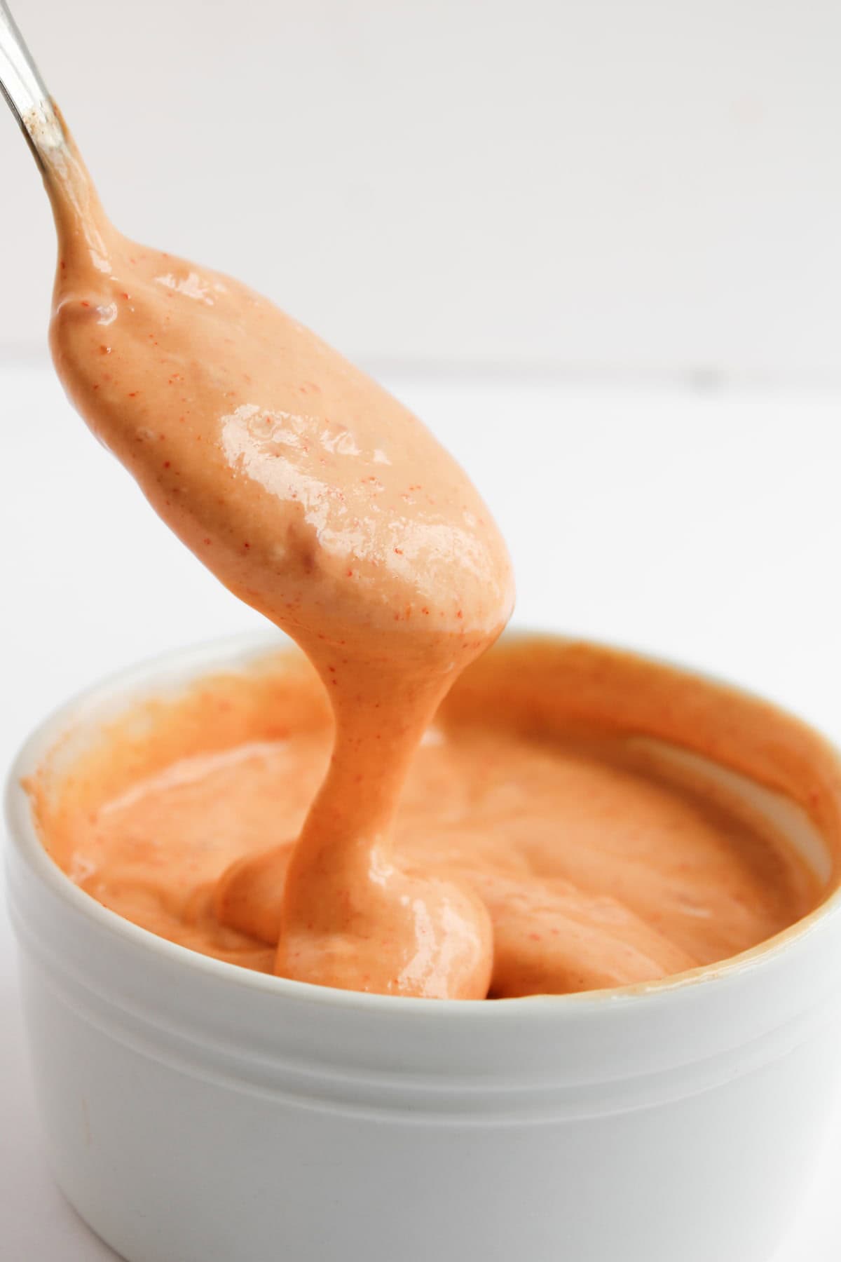 A spoon lifts a thick, orange-pink sauce from a white ramekin against a plain background.