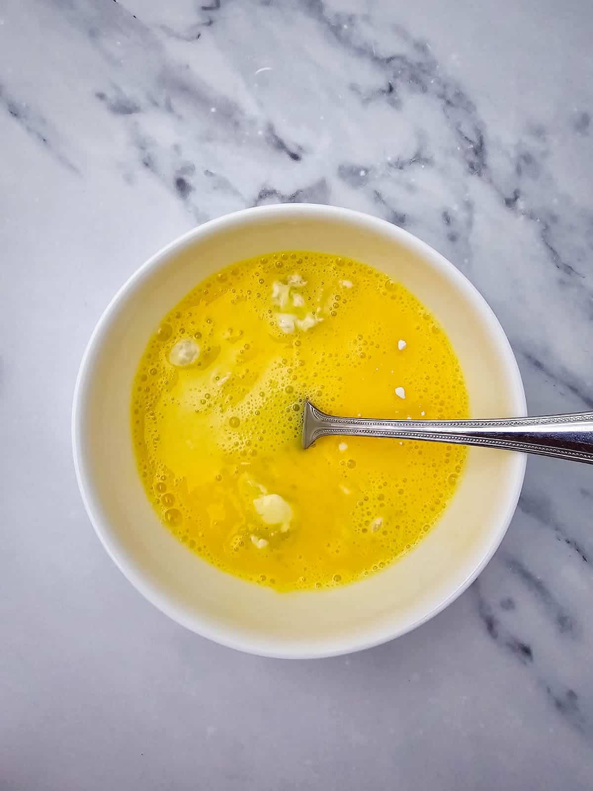 A white bowl containing beaten eggs with small lumps, and a metal fork resting inside, on a light marble surface.