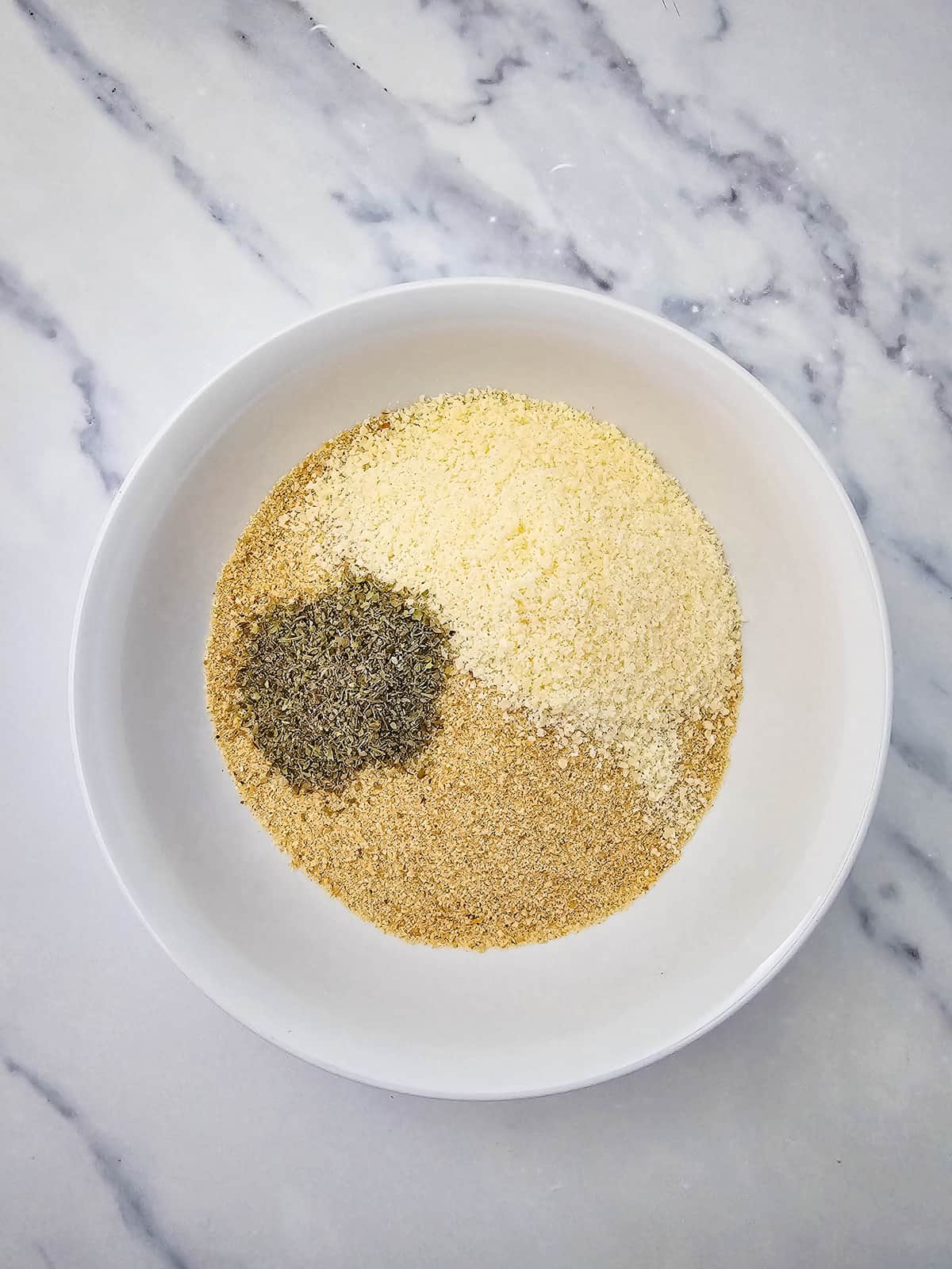 A white bowl on a marble surface containing piles of grated Parmesan cheese, breadcrumbs, and dried herbs.